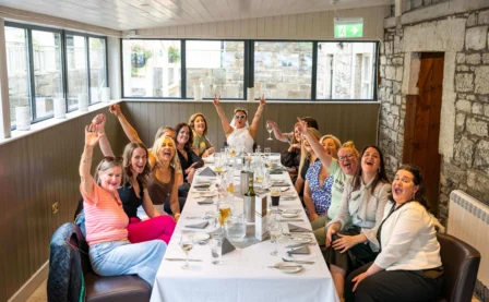 A cheerful group dining in a sunlit rustic room, celebrating with raised arms.