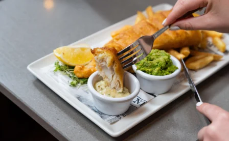 Delicious fish and chips with mushy peas on a table, hands preparing to enjoy.