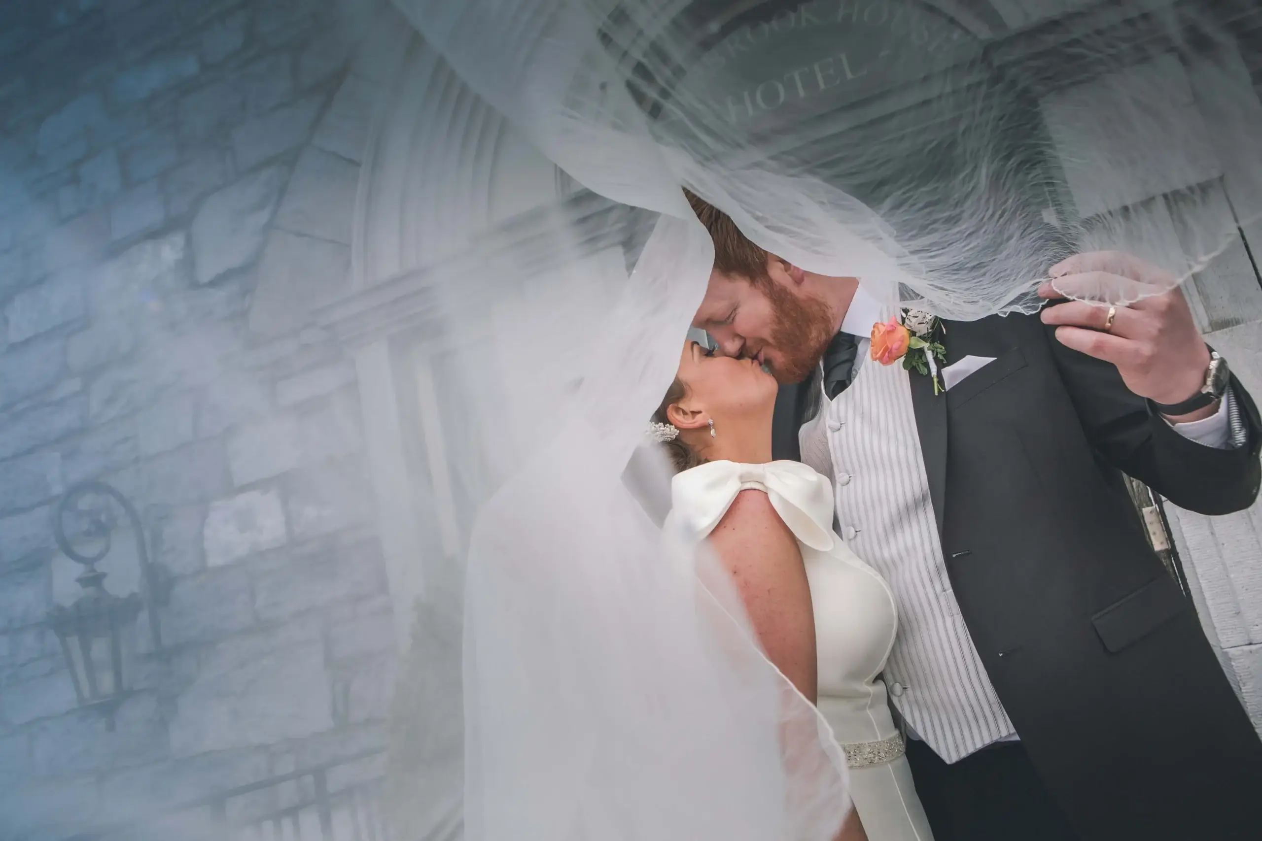 A couple kisses under a bridal veil at a hotel entrance, signifying romance and celebration.
