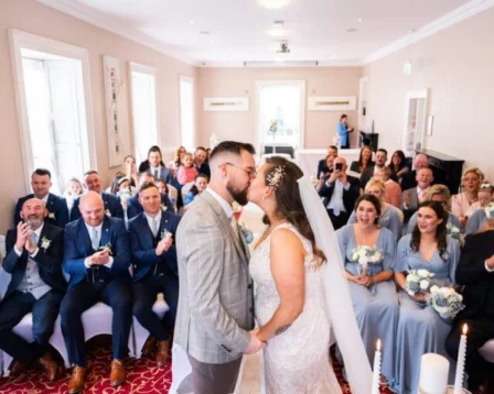 A couple shares a kiss at their wedding ceremony surrounded by family and friends in a cosy, elegant room.