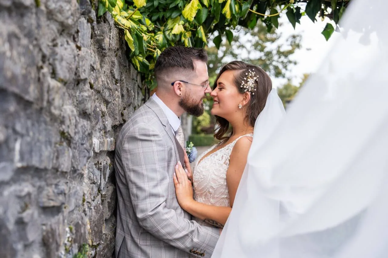 A joyful couple embraces by a rustic stone wall, surrounded by lush greenery.
