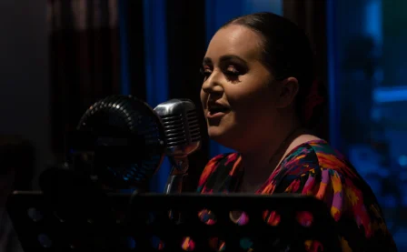 A woman sings warmly into a vintage microphone in a cosy, dimly lit room.