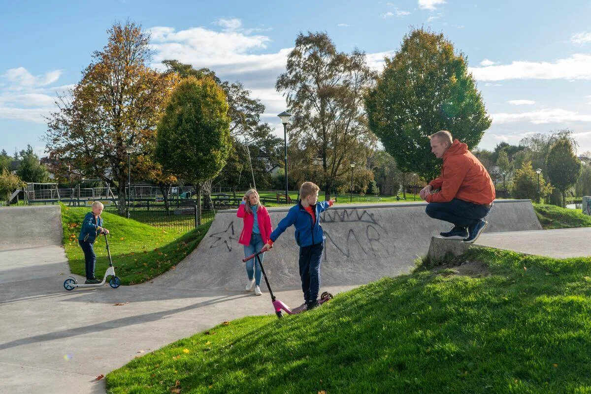A family enjoys scootering in a bright, green park on a sunny day.