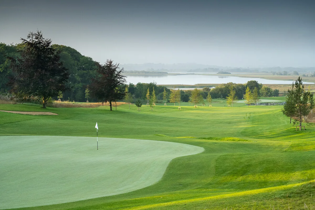 A scenic golf course with rolling green hills near a tranquil lake under a grey sky.