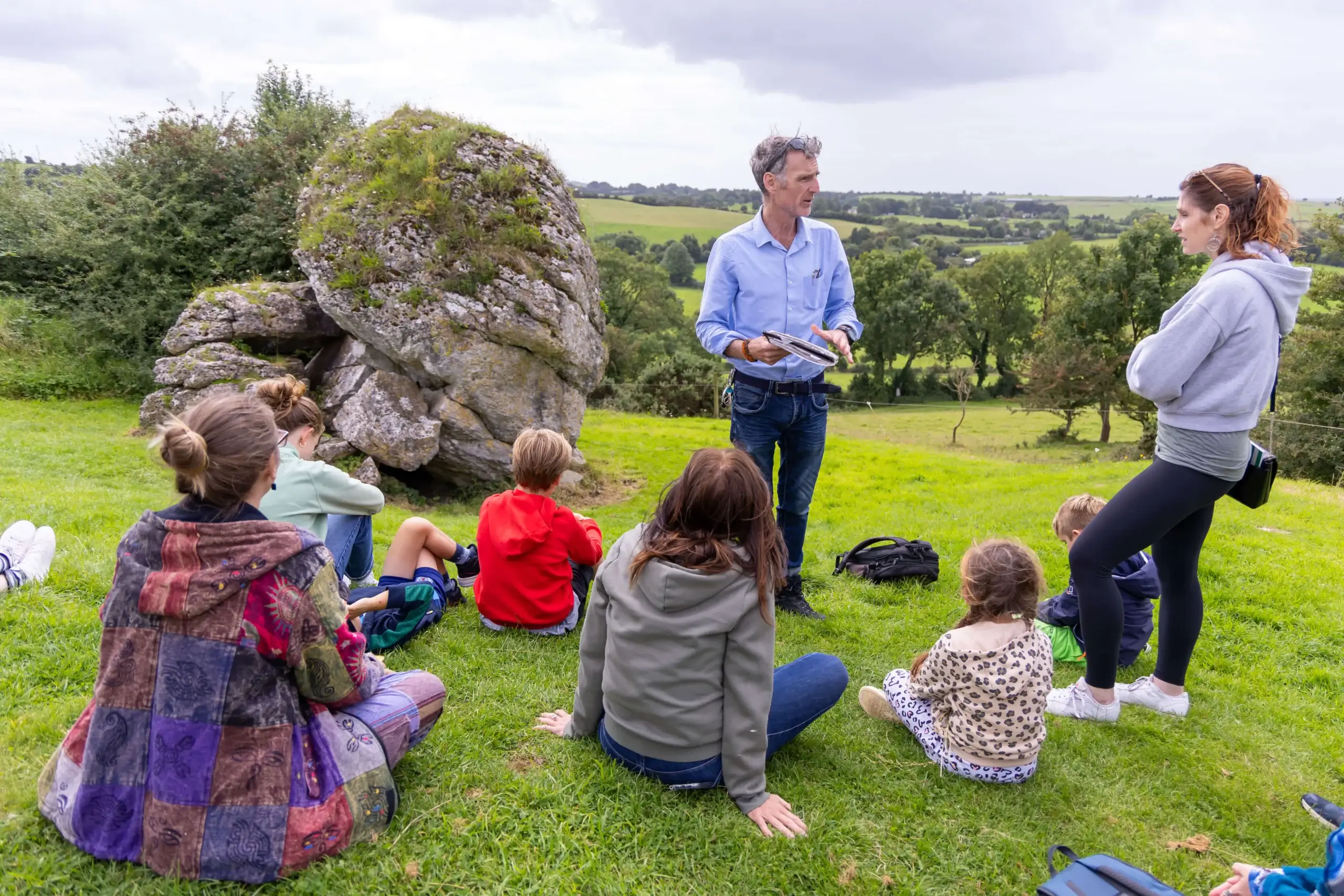 Family listens to guide near scenic rock in lush countryside setting.