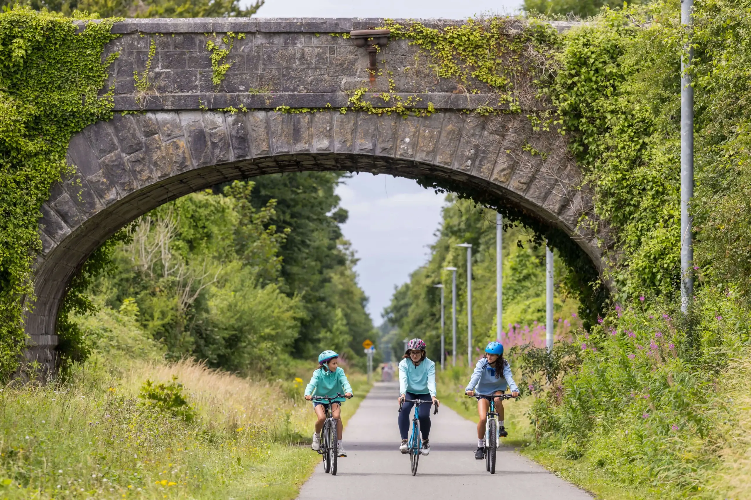 Family cycling under a stone bridge in lush, green countryside.