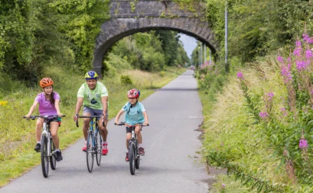 Family cycling on a scenic country path under a stone bridge, surrounded by greenery.