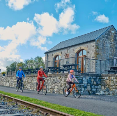 Family cycling by a rustic stone building under a bright blue sky.