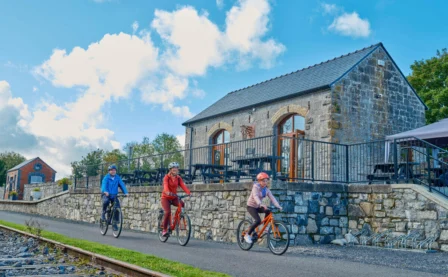 Family cycling by a rustic stone building under a bright blue sky.