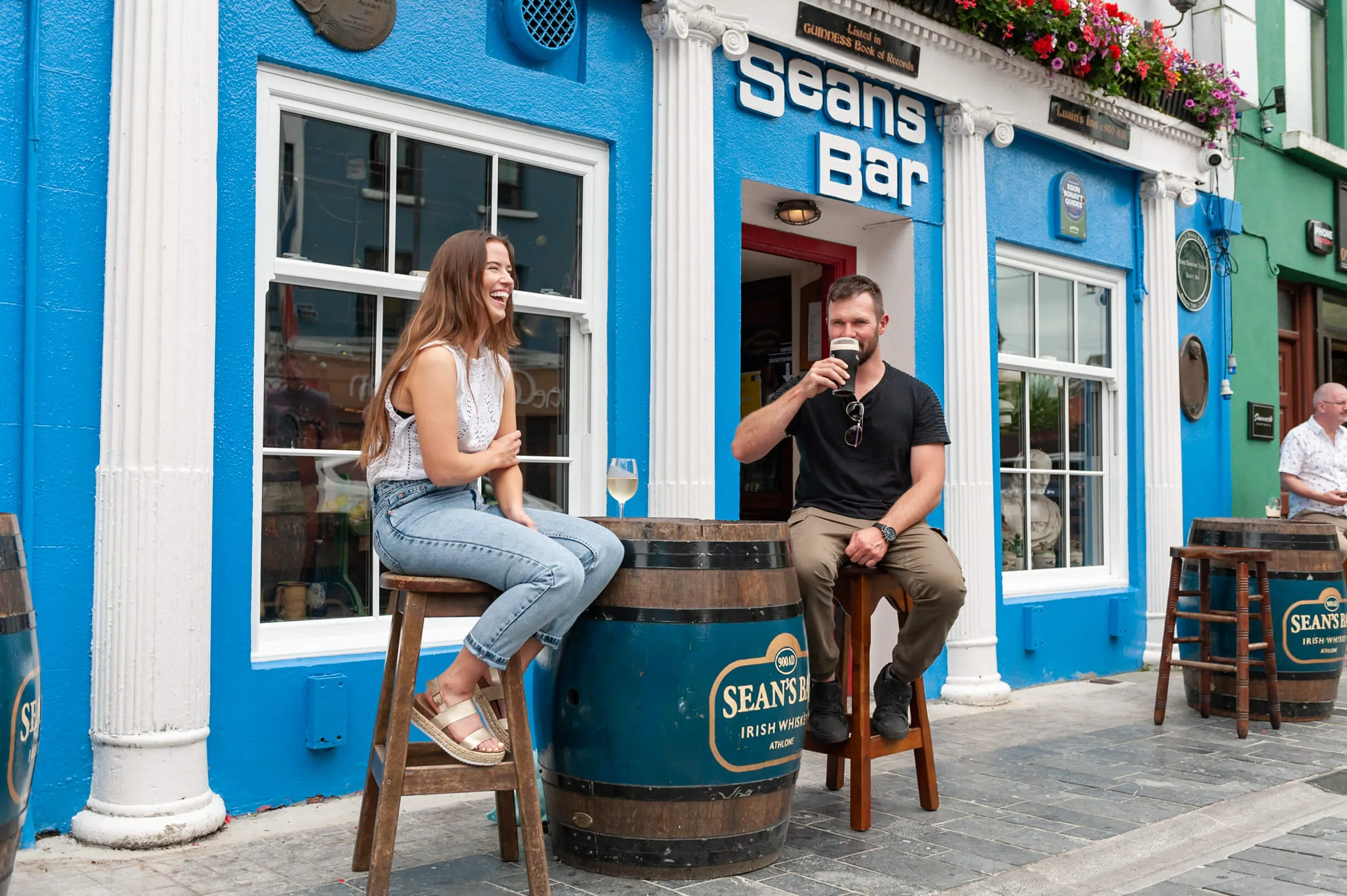 A smiling couple enjoys drinks at a vibrant, blue-painted pub with colourful floral displays.