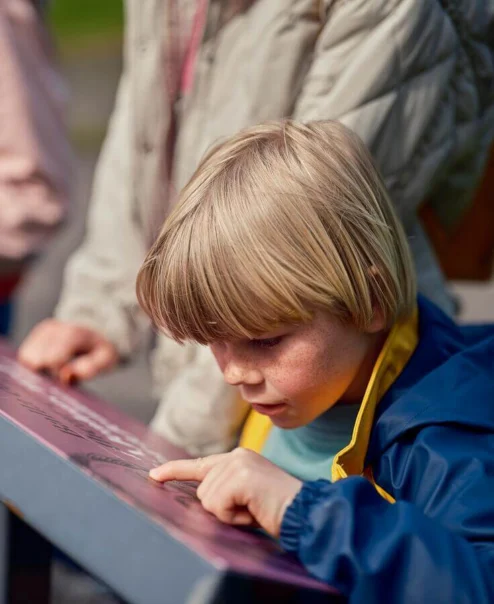 A young child points at a sign outdoors with family nearby.
