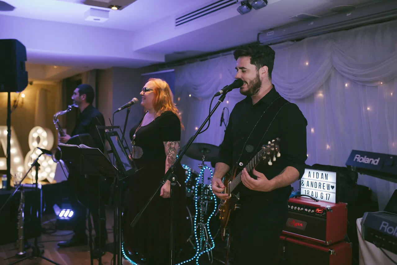 A band plays lively music at a cosy hotel venue, featuring a saxophonist, guitarist, and vocalist.