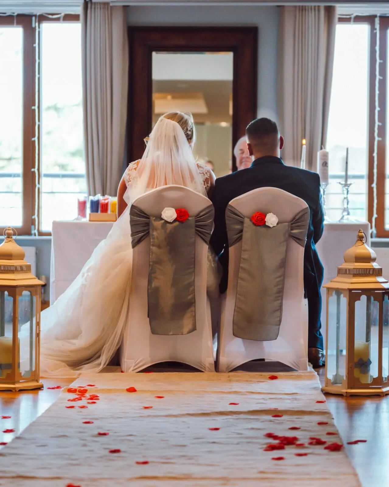 A couple sits during a wedding ceremony in a bright, elegantly decorated room.