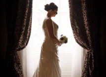 A bride in an elegant white gown stands by a window with lavish curtains, holding a bouquet.