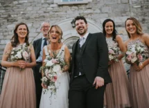 A joyful wedding party in front of a stone venue, the couple and bridesmaids are smiling warmly.