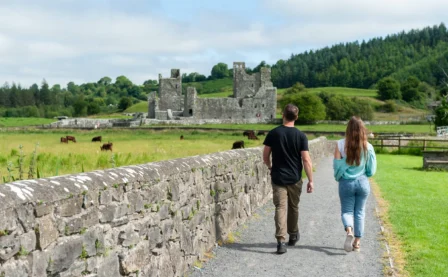 A couple strolls towards an ancient castle, surrounded by lush greenery and grazing cows.