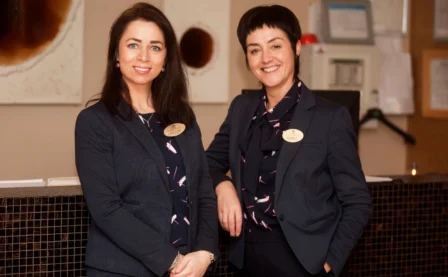 Two smiling hotel receptionists in uniforms standing at the front desk, ready to assist guests.