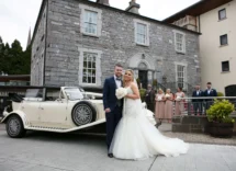 A couple poses in front of a classic car at a charming stone hotel with family nearby.