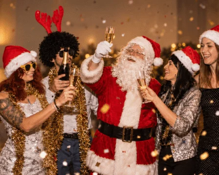 Festive group celebrating with Santa, wearing holiday hats, toasting champagne in a cosy setting.