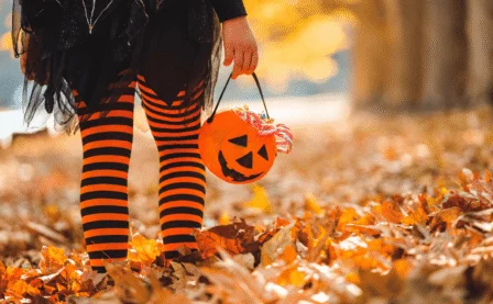 Child in Halloween costume holding a pumpkin bucket, standing on autumn leaves.