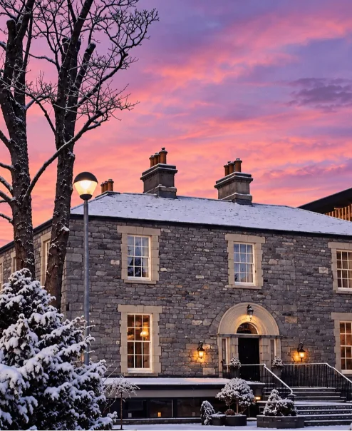 A historic stone hotel warmly lit at sunset, surrounded by snow, welcoming guests in winter.