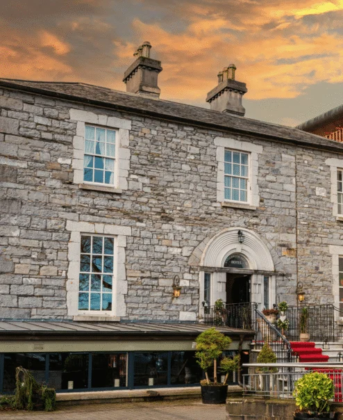 Historic stone hotel with warm sunset, lush trees, and inviting entrance with red carpet.