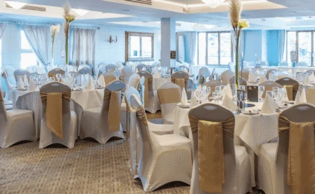 Elegantly dressed banquet room with round tables set for a family celebration, white linens, gold chair sashes, glassware and folded napkins — bright windows visible