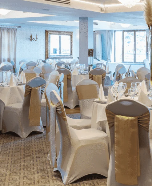 Elegantly dressed banquet room with round tables set for a family celebration, white linens, gold chair sashes, glassware and folded napkins — bright windows visible